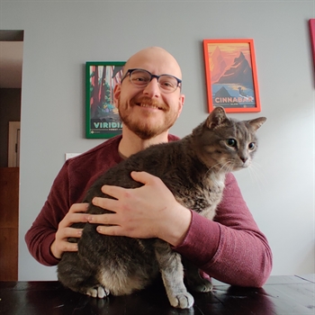 Dan Matthews wears glasses, smiles, and embraces a beautiful cat