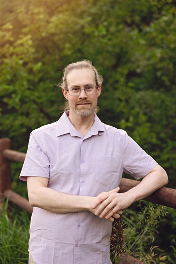 Image of a man with glasses, Joshua Newport, leaning against a fence with trees in the background.